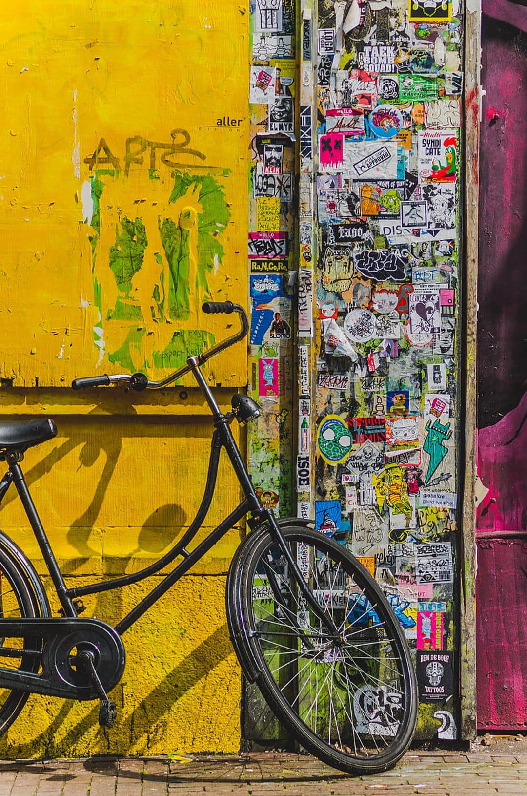 Black vintage bicycle leaning against a bright yellow wall covered in colorful stickers and urban graffiti tags.