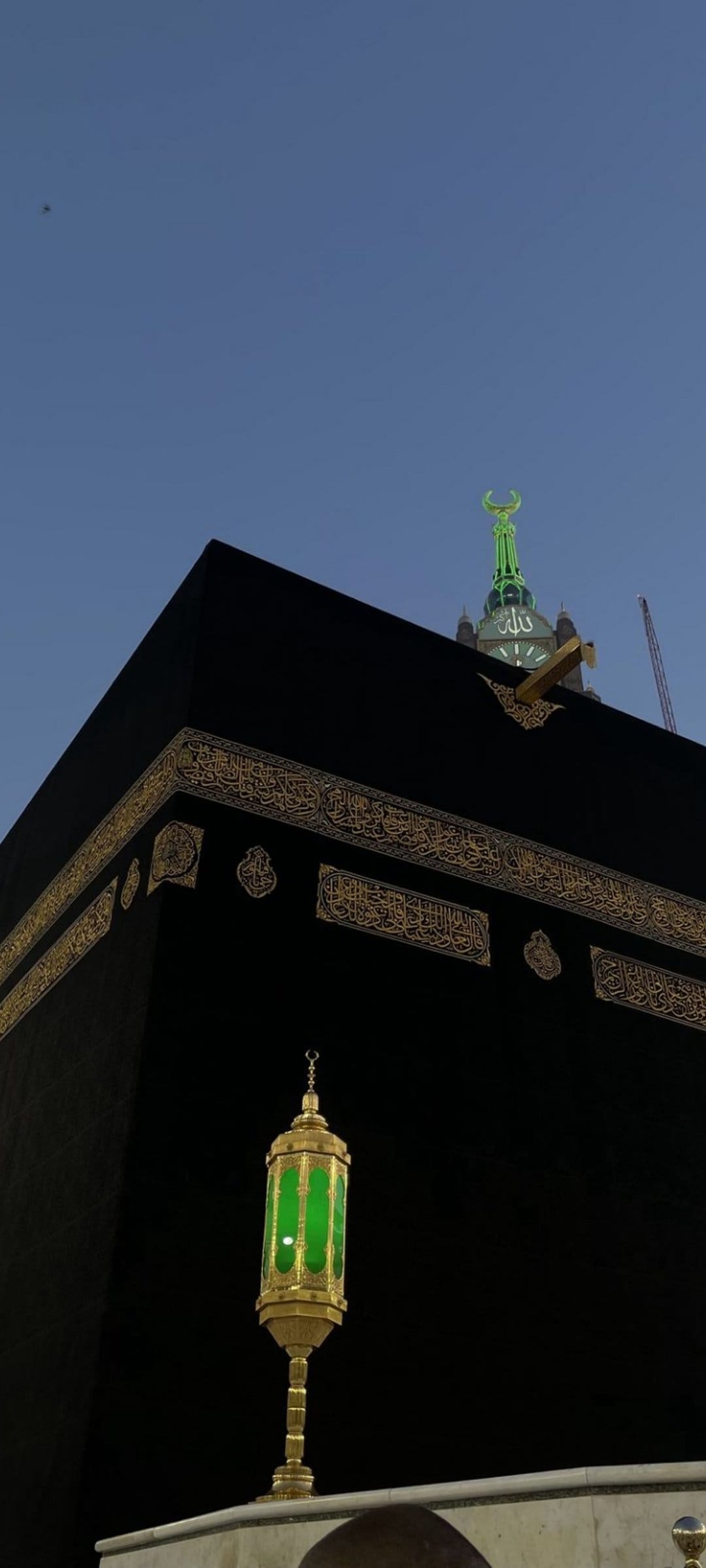 The Kaaba with gold calligraphy stands behind a green glass lantern under a crescent moon spire against a blue sky.