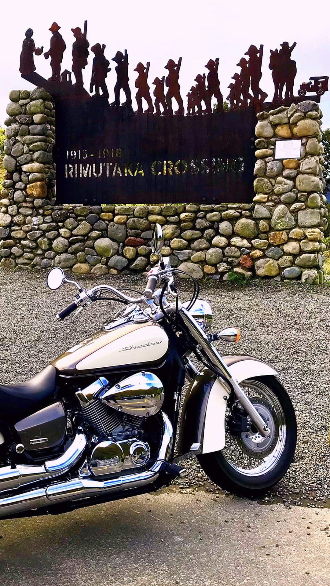 Cream motorcycle parked by a stone monument featuring metal soldier silhouettes under a bright outdoor sky.