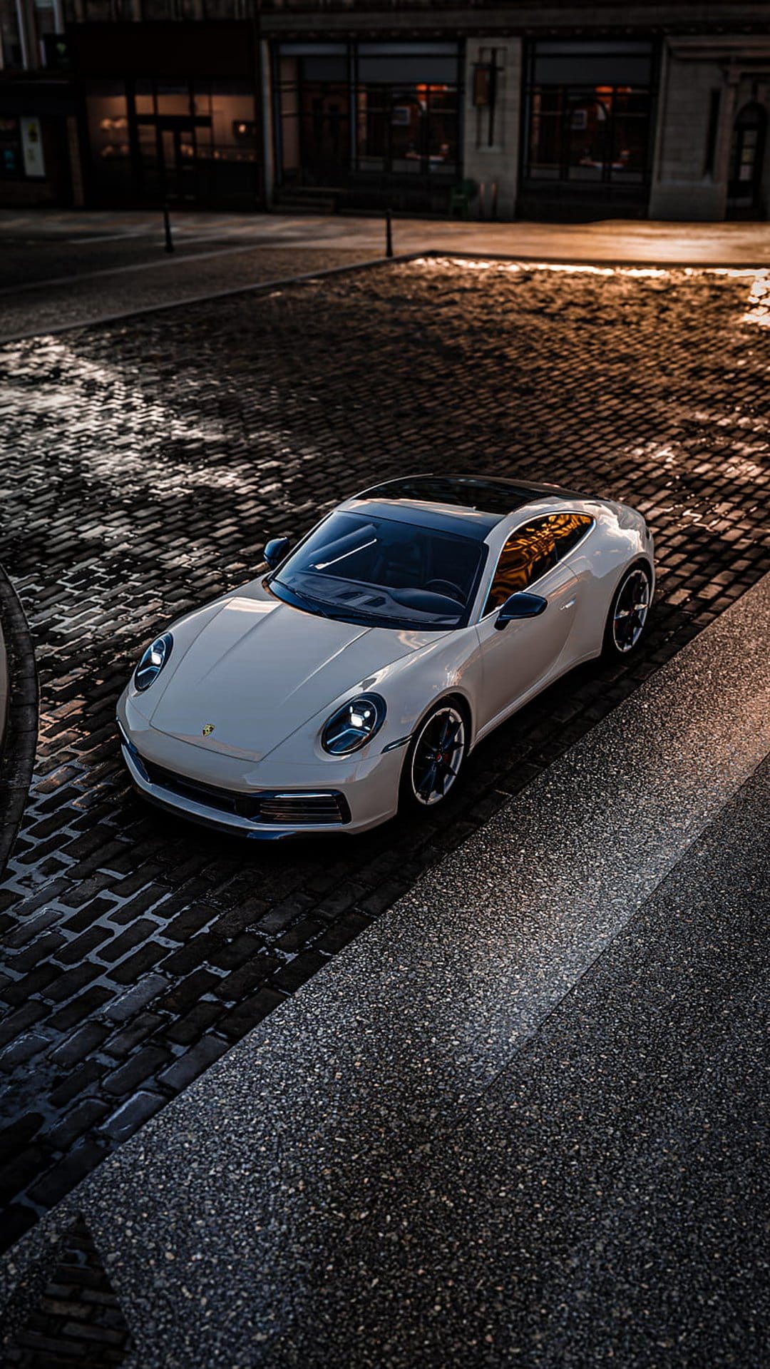 White Porsche 911 parked on wet cobblestones at dusk with warm streetlights reflecting on the sleek car body.