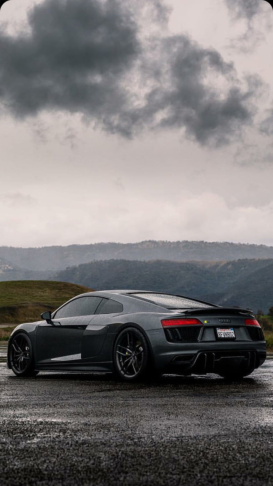 Audi R8 supercar parked on a wet asphalt road under a dramatic, cloudy sky with distant hills and reflections.