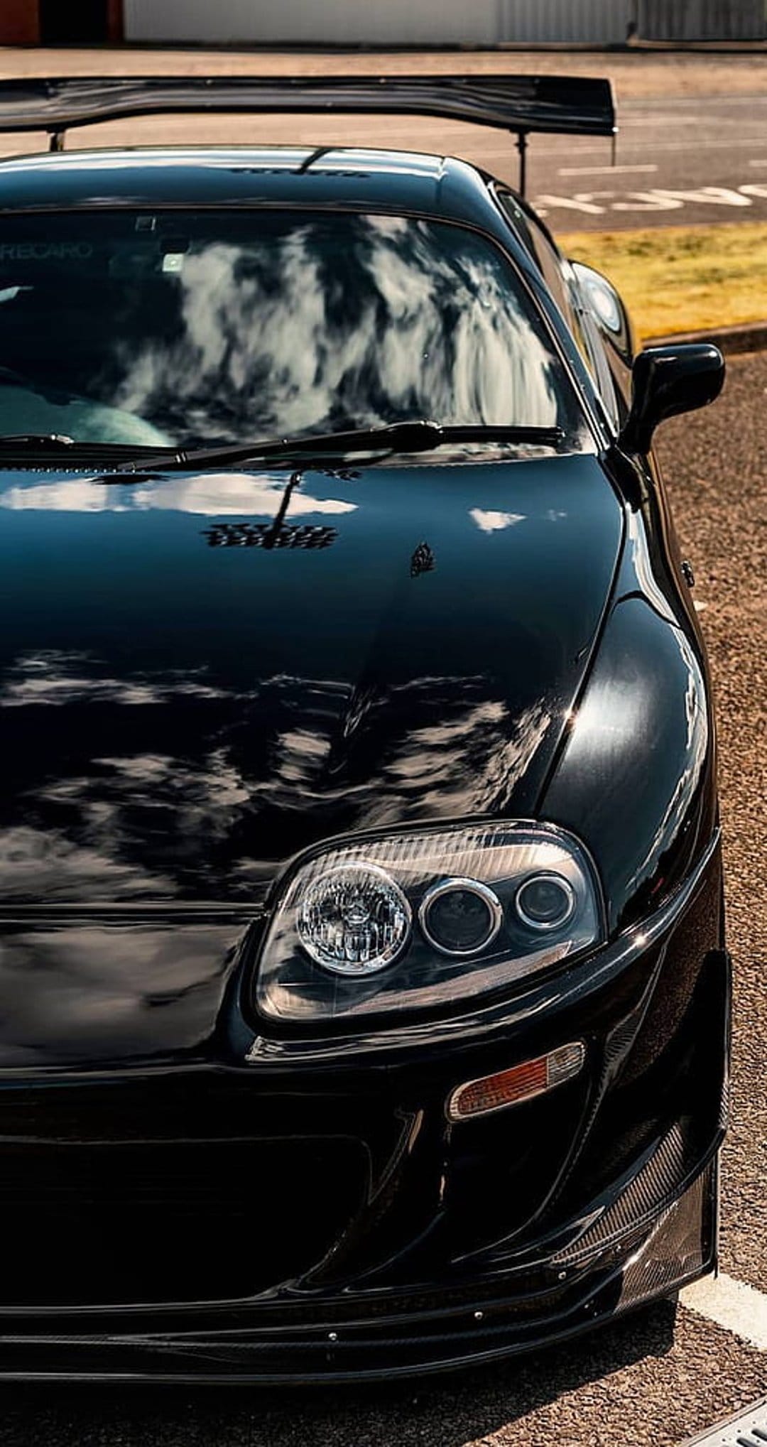 Black Toyota Supra with a large rear wing reflects white clouds on its glossy hood and windshield in daylight.