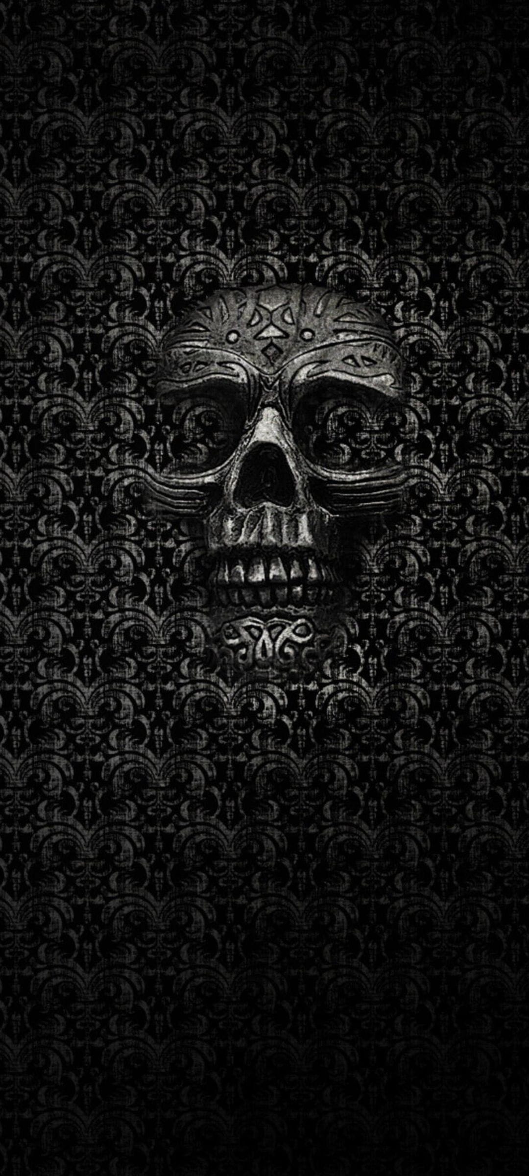 Human skull with intricate floral engravings centered against a dark background of repeating filigree patterns.