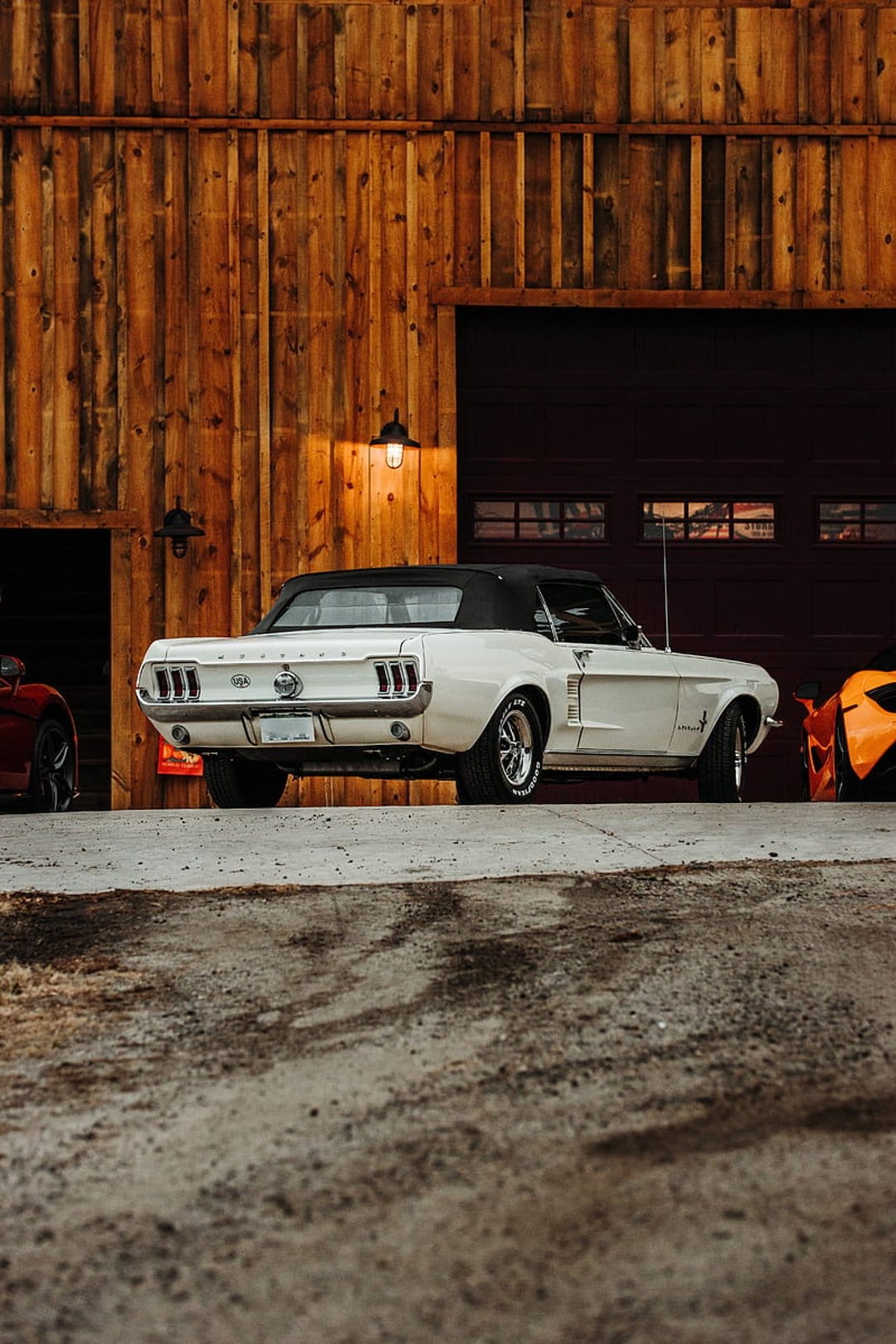 White Ford Mustang convertible parked in front of a weathered wooden garage with an orange car visible nearby.