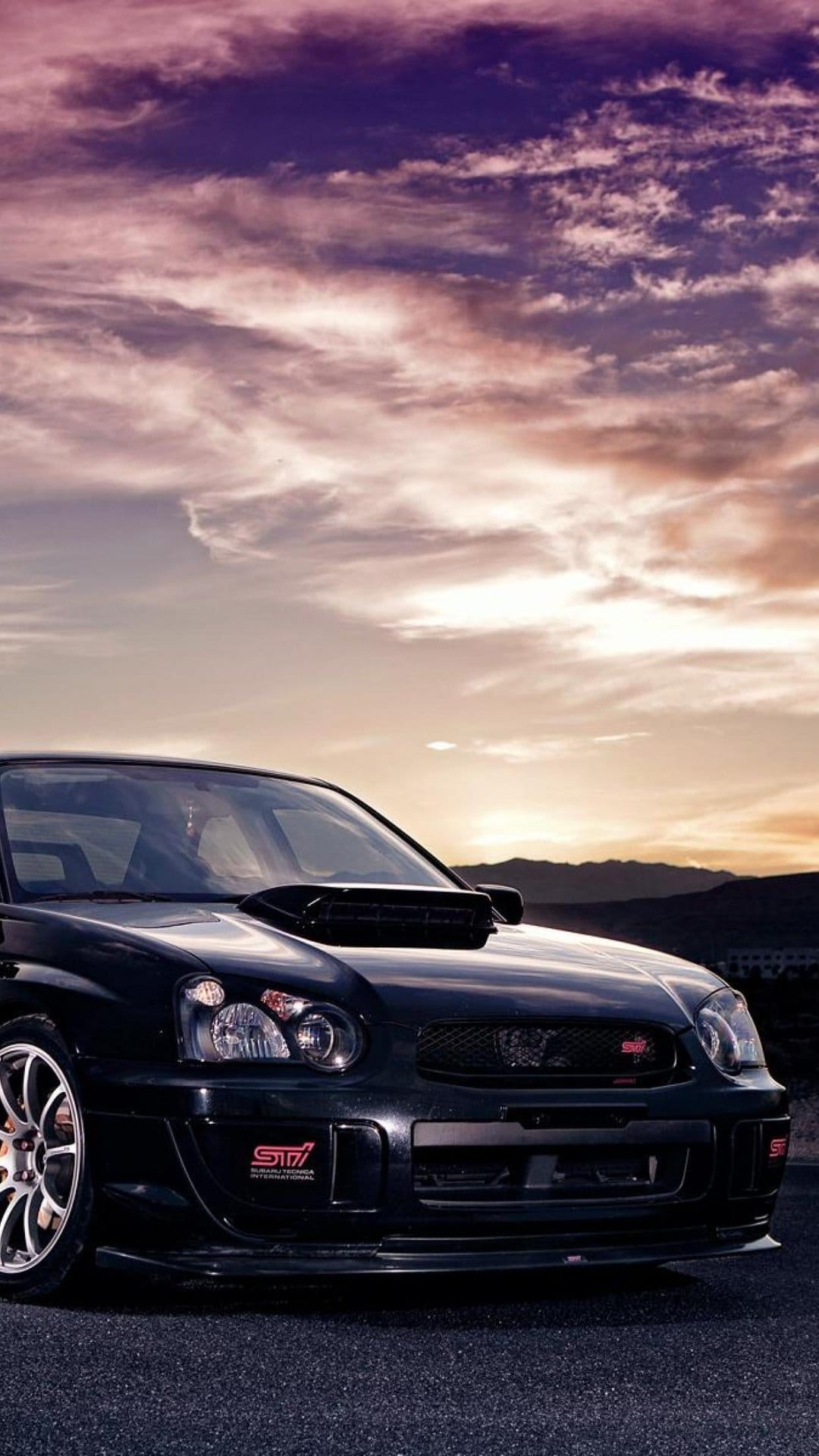 Black Subaru WRX STI with a prominent hood scoop parked under a dramatic purple and orange sunset sky.