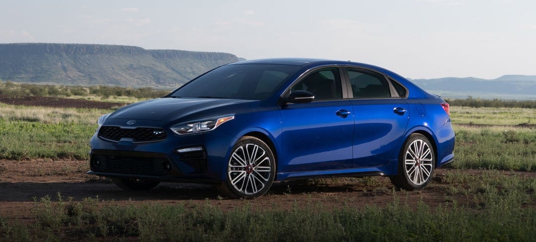 Blue Kia Forte GT sedan on a dirt path with distant plateaus under a clear sky in a wide desert landscape.