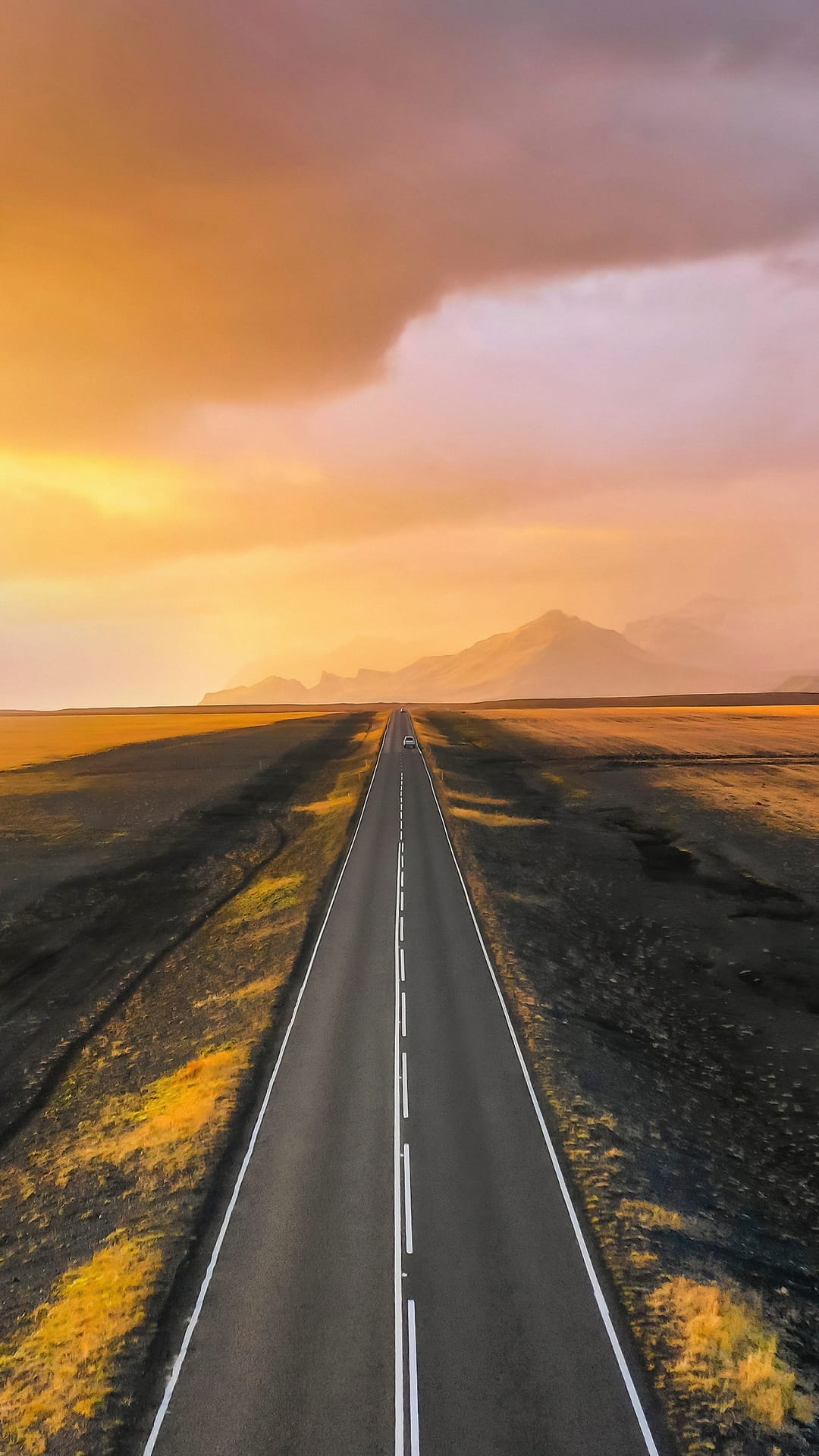 Straight asphalt road stretching through a desert toward distant mountains under a warm orange sunrise sky.