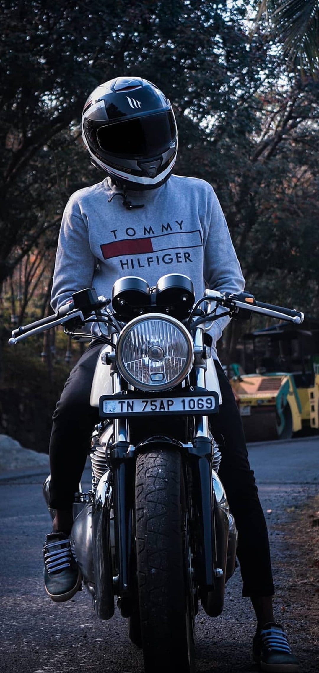 Man in a black helmet and grey sweatshirt sits on a motorcycle with a clear headlight and yellow vehicle behind.