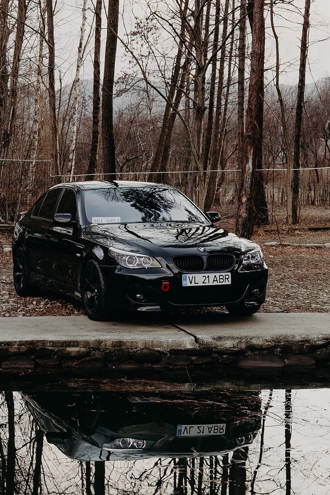 Black BMW sedan parked on a concrete platform beside a reflective lake with a dense forest in the background.