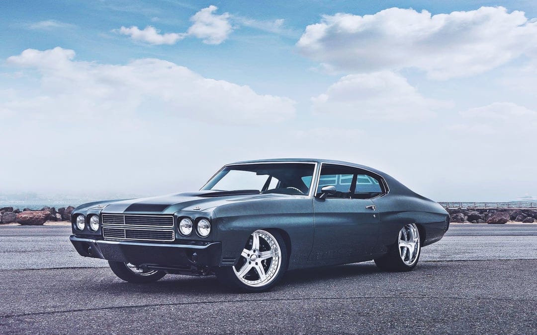A dark grey 1970 Chevelle muscle car with chrome rims parked on asphalt under a bright cloudy sky.