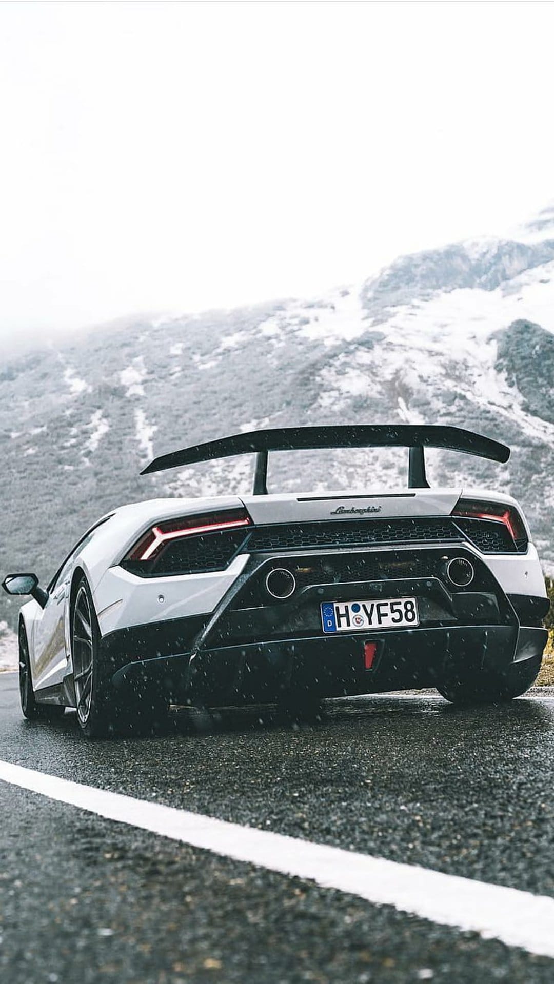 White Lamborghini Huracan Evo RWD Spyder with a rear wing driving on a wet road amidst snowy mountains and mist.