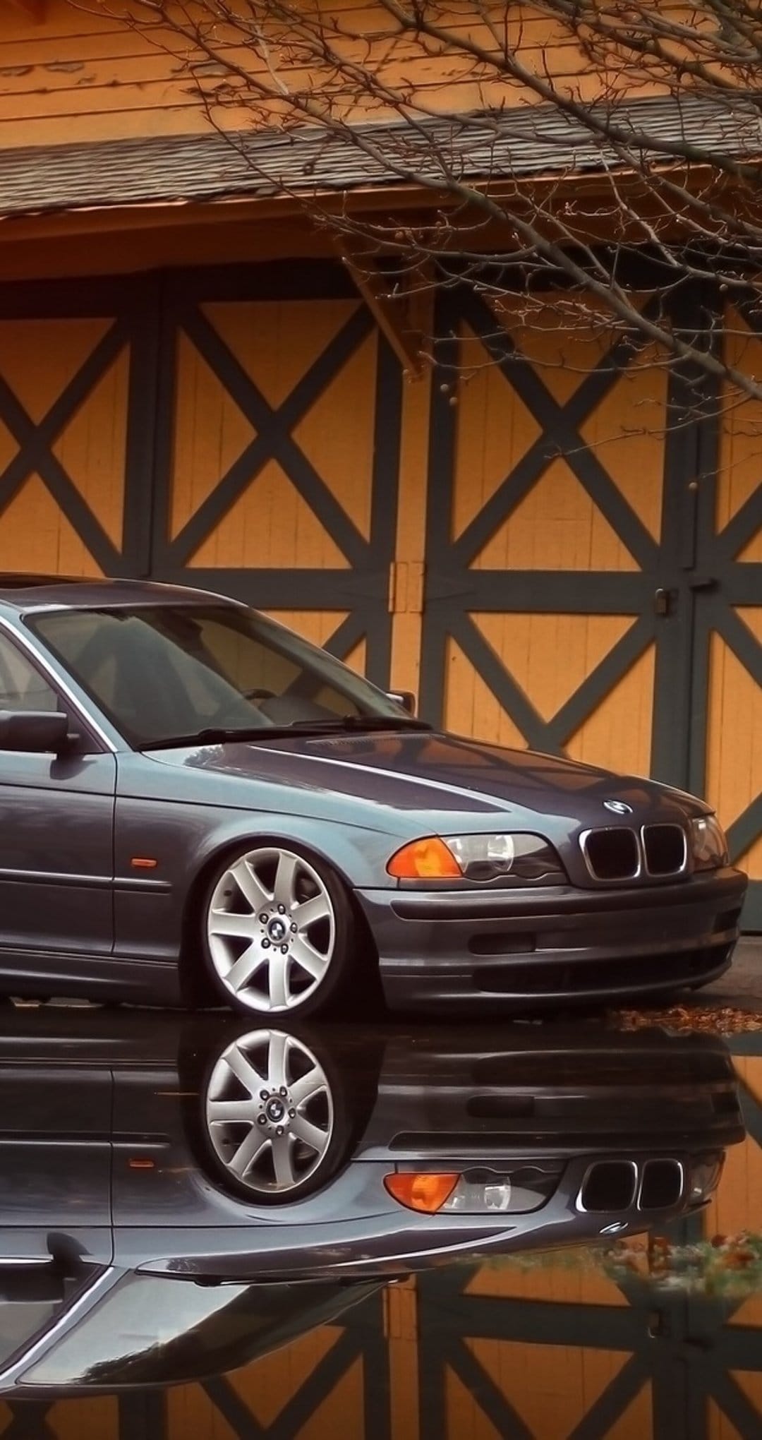 Charcoal BMW sedan reflecting on a wet ground in front of weathered orange barn doors and bare branches.