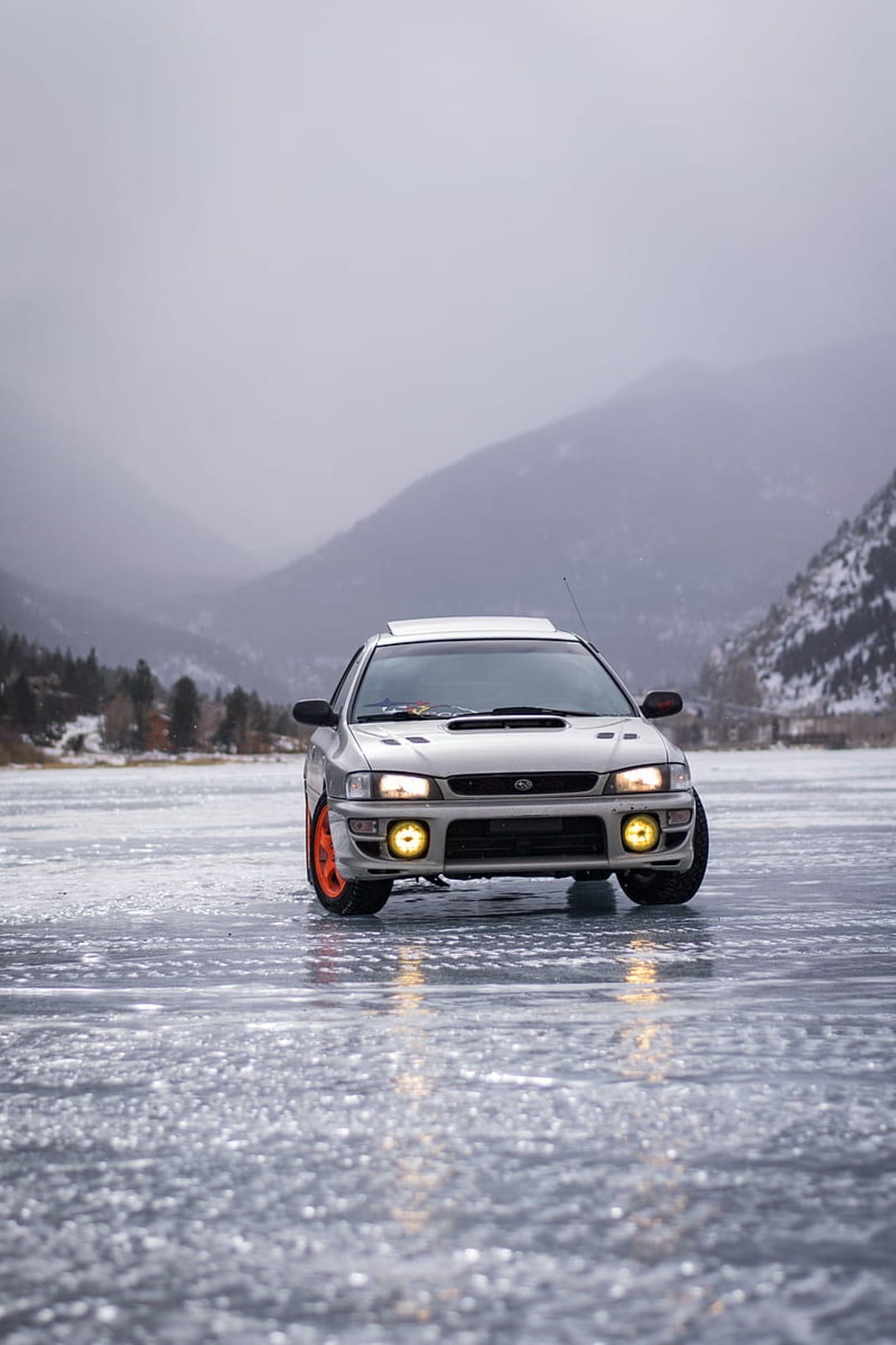 Silver Subaru Impreza with orange wheels parked on a cracked frozen lake under a dramatic mountain sky backdrop.