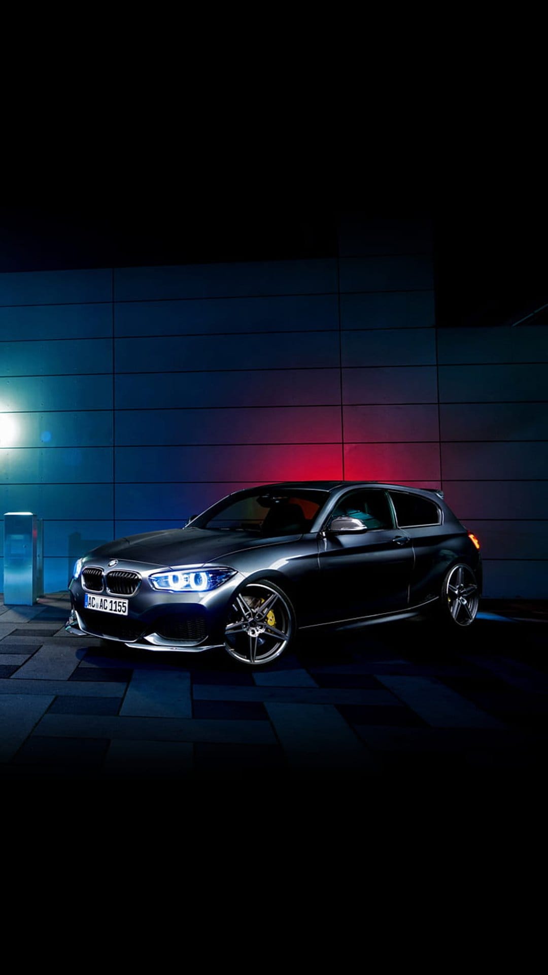 Dark BMW hatchback with glowing headlights parked near a building under blue and red neon ambient lighting.