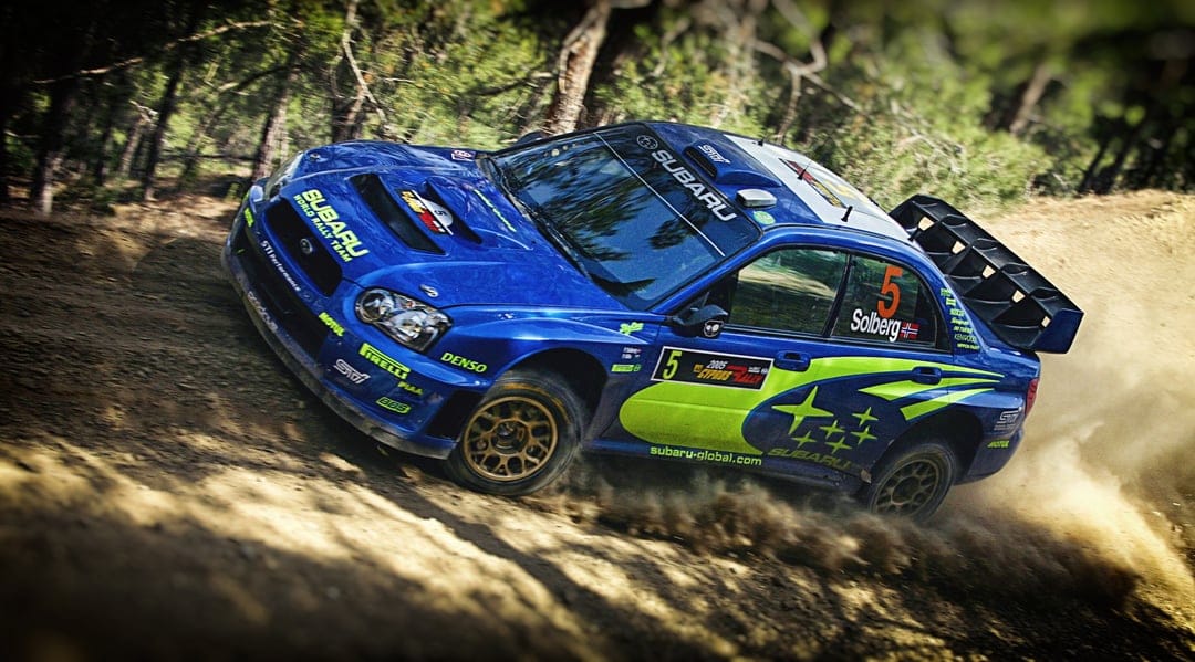 Blue Subaru rally car drifting through a forest trail kicking up a large cloud of golden dust.