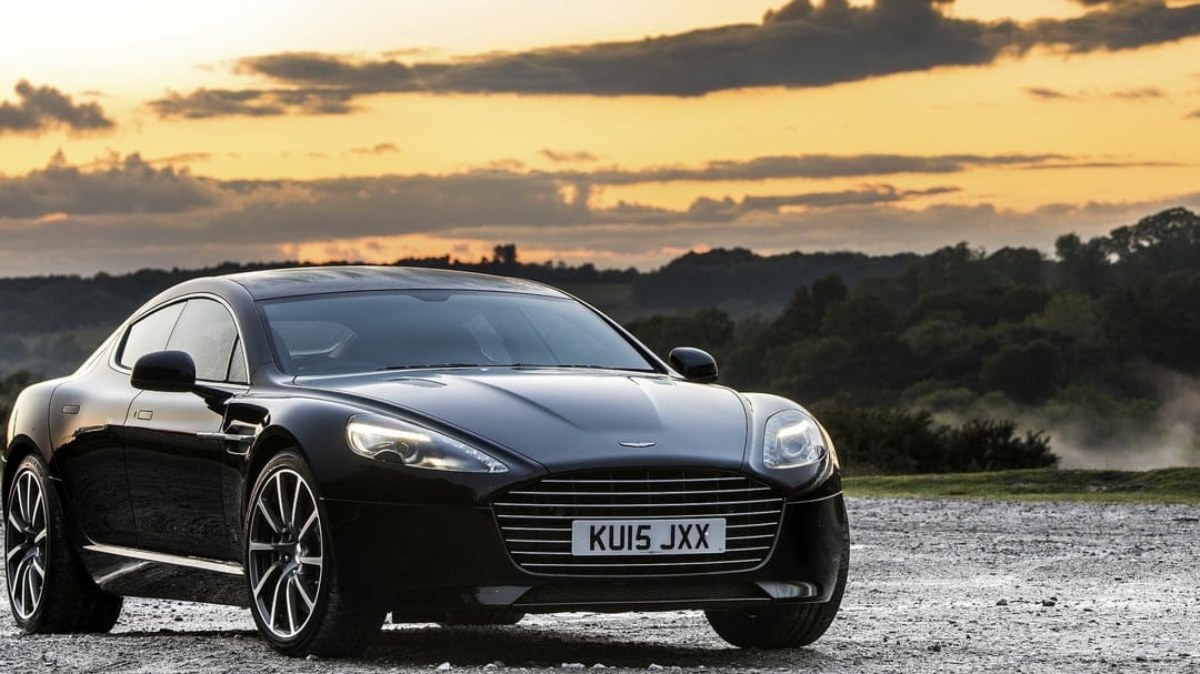 Black Aston Martin Rapide S parked on gravel under a golden sunset sky with distant green hills and trees.