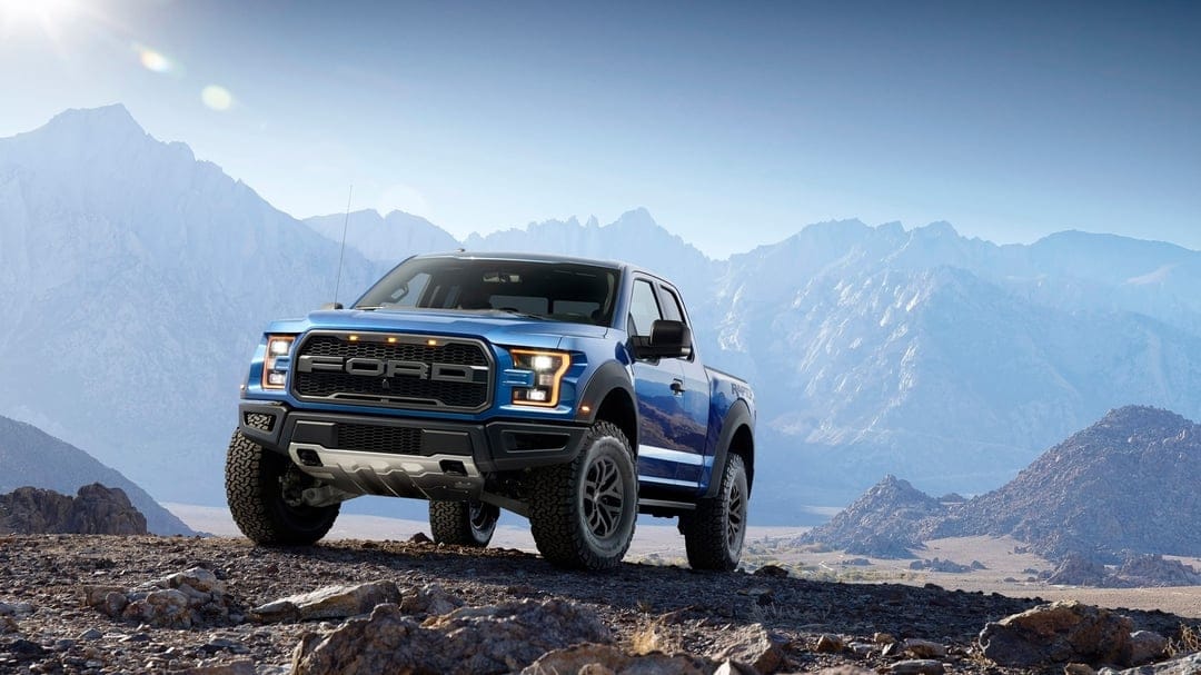 Blue Ford Raptor truck parked on a rocky hill with sharp snow-capped mountains and a clear sky behind it.