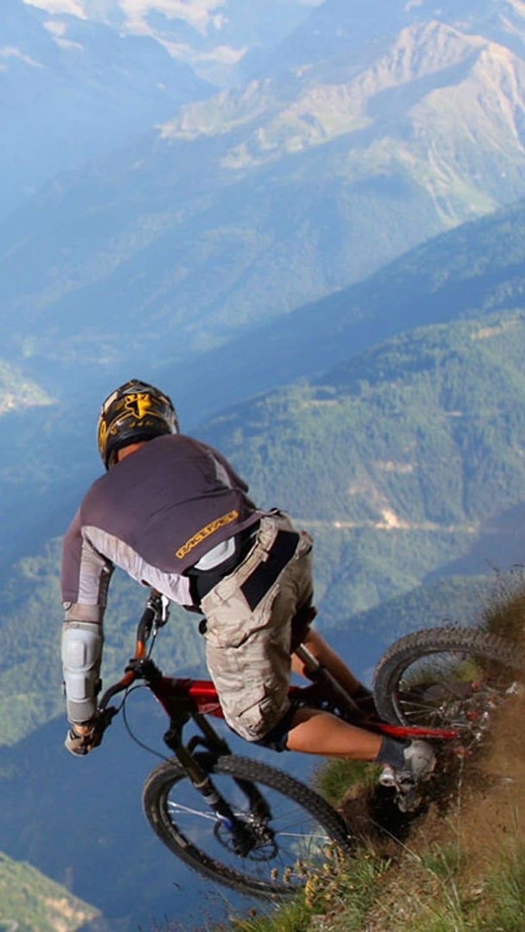 Mountain biker in protective gear leans forward while riding down a steep, rocky trail toward misty blue peaks.