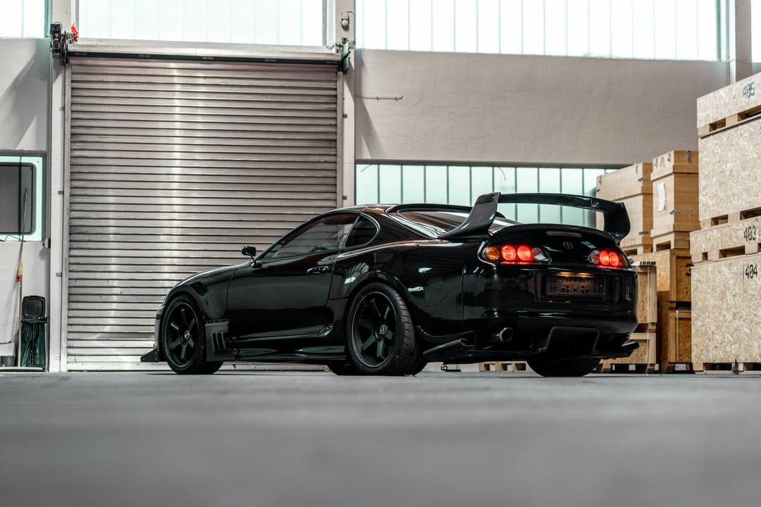 Black Toyota Supra with a large rear spoiler parked in a warehouse next to wooden crates and a garage door.