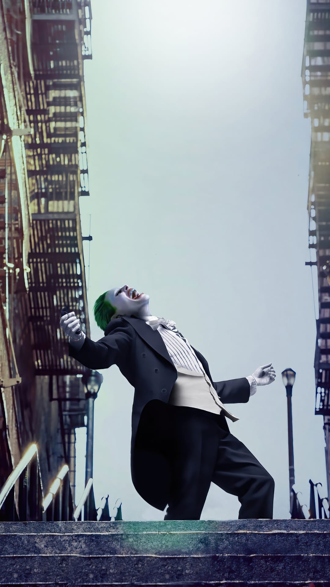 Joker with green hair laughs on stone stairs between city buildings wearing a black suit and striped vest.