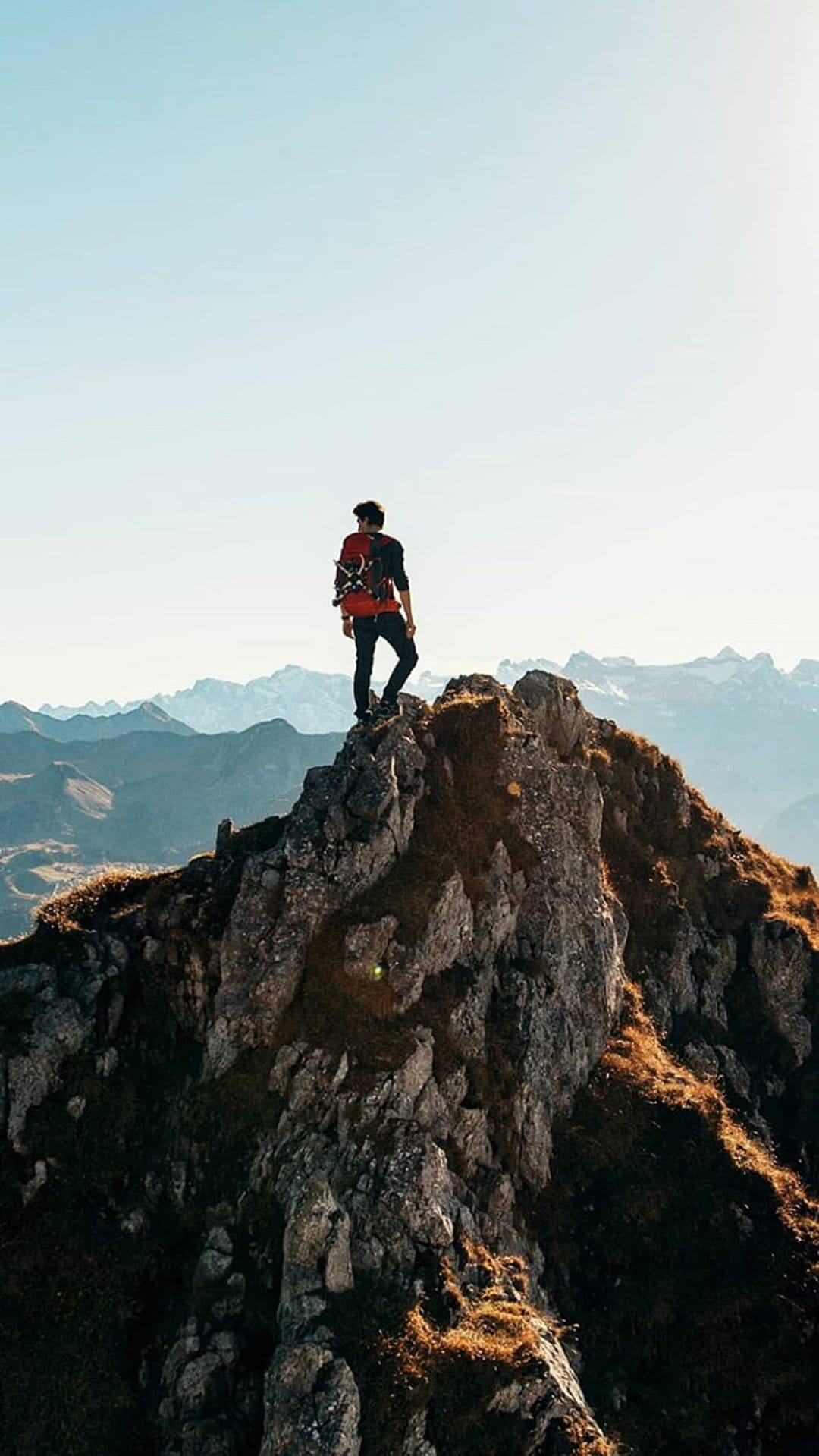 Hiker with a red backpack stands on a rocky summit facing distant snow-capped mountains and a clear blue sky.