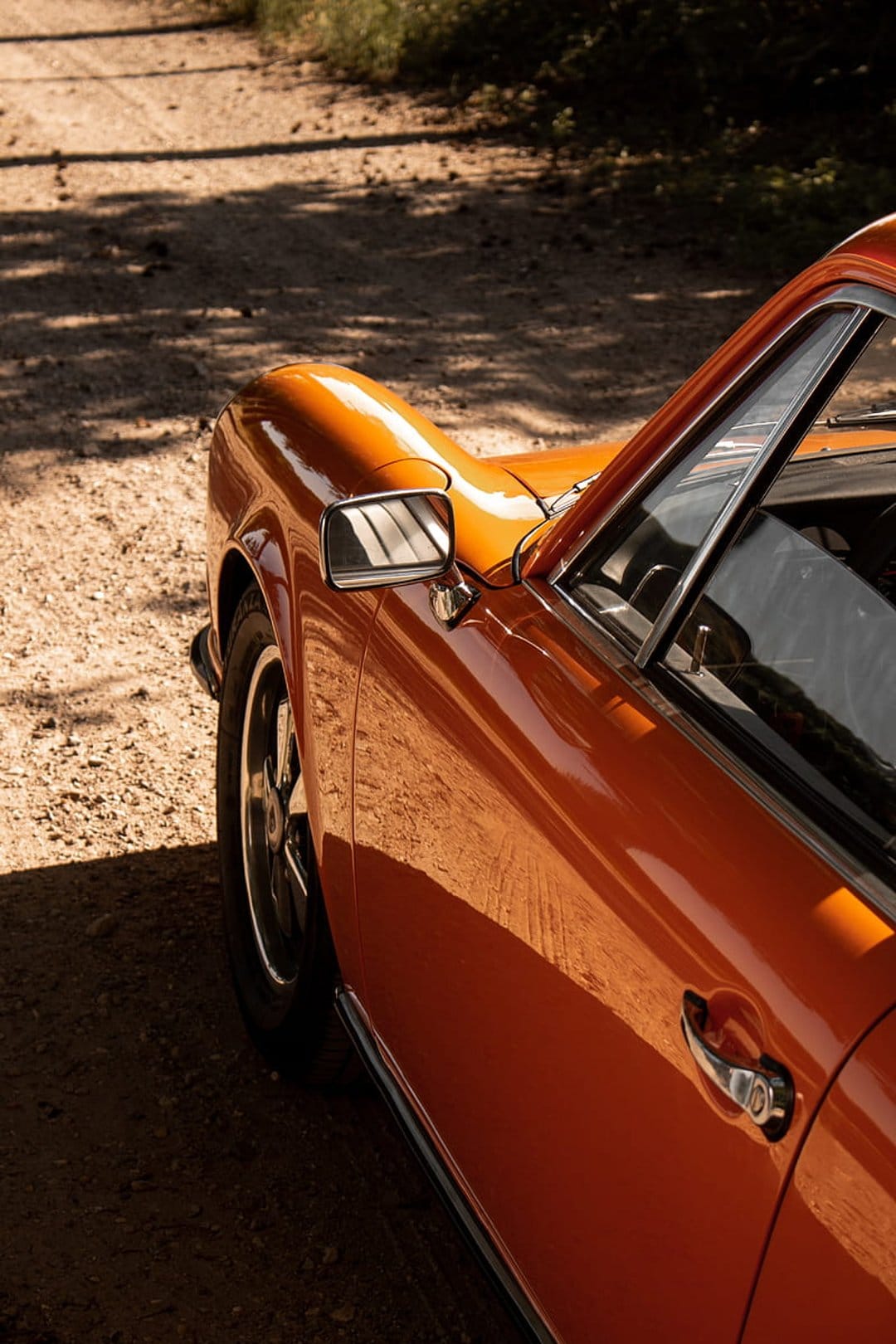 Orange vintage Porsche coupe parked on a dusty dirt road with sunlight reflecting off the polished side mirror.
