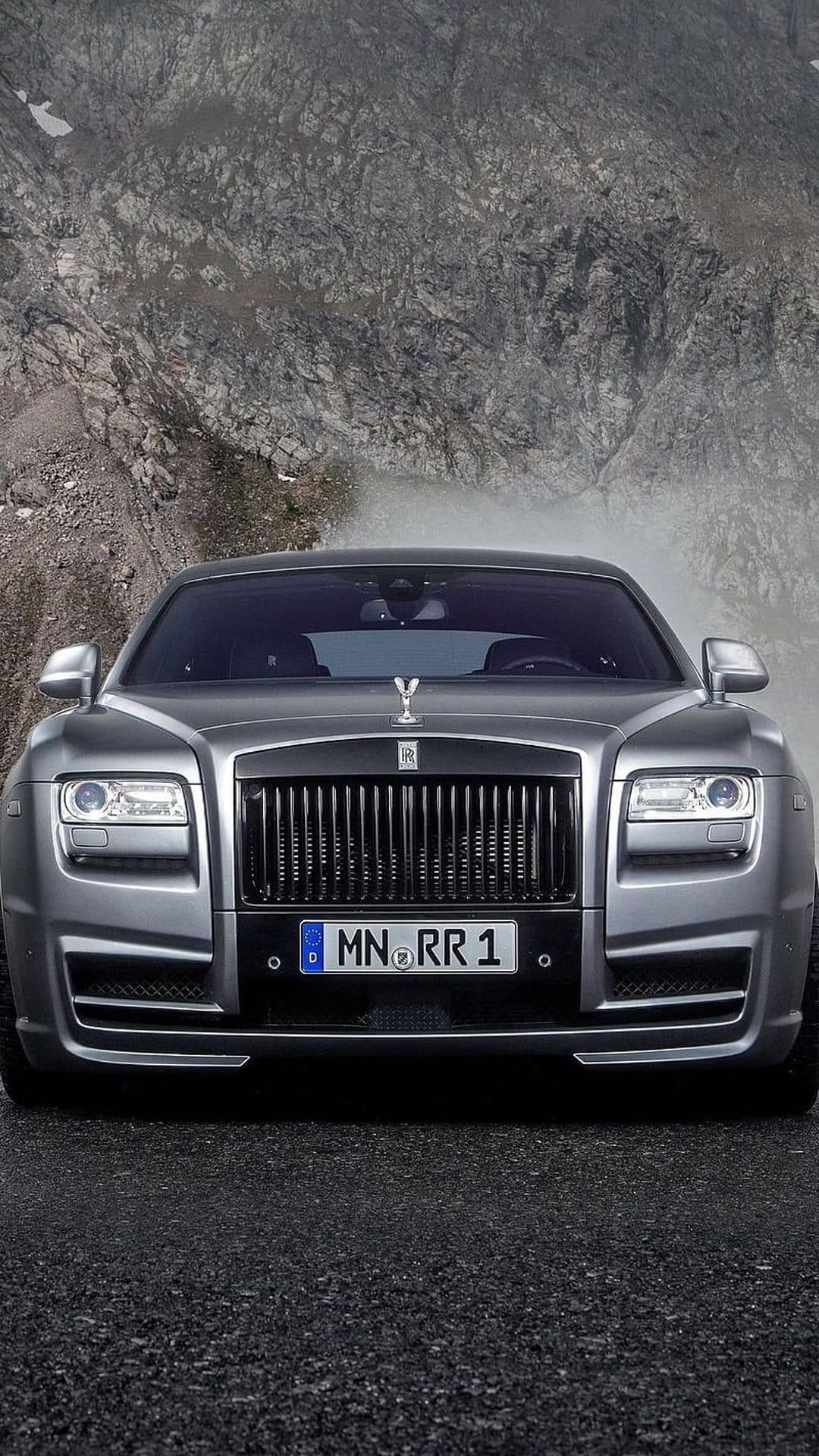 Silver Rolls-Royce Ghost parked on wet asphalt before a jagged, misty mountain range under a cloudy sky.