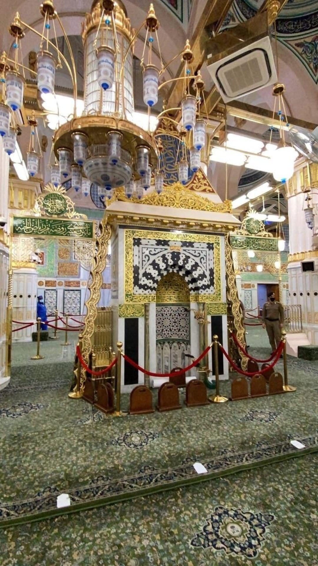 Large tiered chandelier hanging over a green patterned carpet inside a mosque with golden calligraphy on walls.