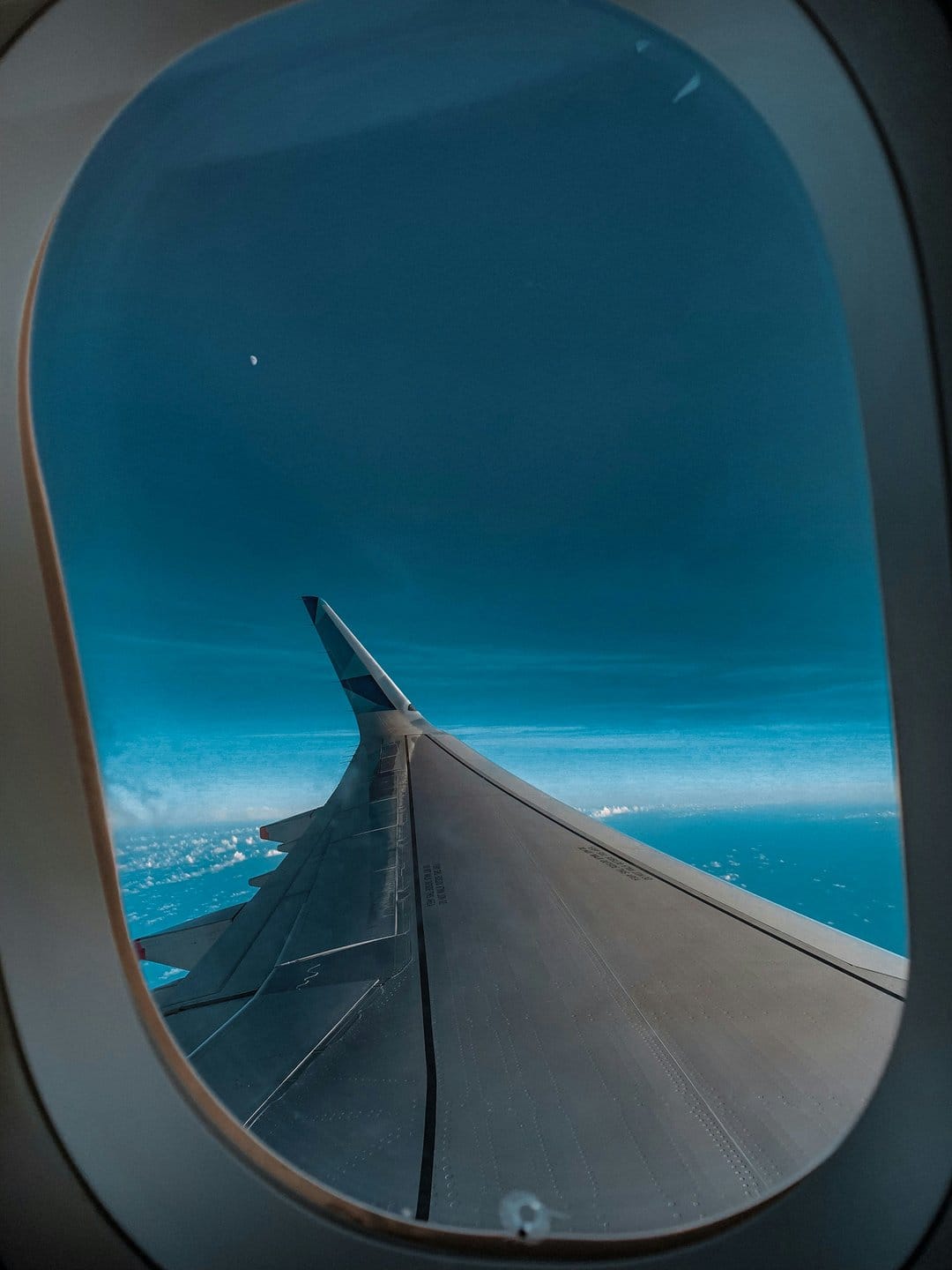 Airplane wing extending over white cumulus clouds and the deep blue ocean from a high altitude window perspective.