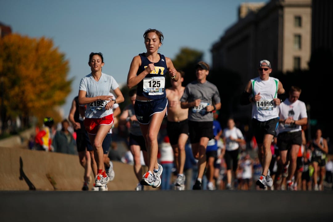 Marathon runners with bib number 125 stride past autumn trees on a sunlit city street during a road race.