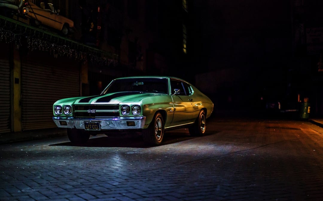 Green classic muscle car with racing stripes parked on a dark cobblestone street under dramatic night lighting.