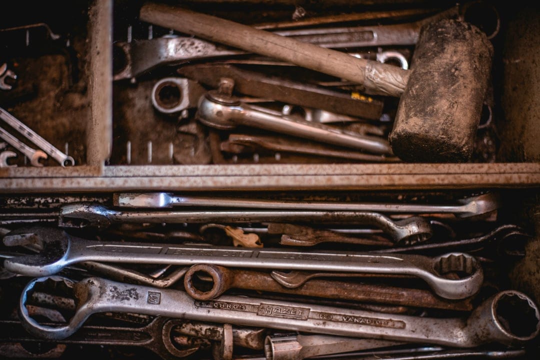 Weathered steel wrenches and a heavy sledgehammer rest in a worn metal toolbox with visible rust and grease marks.