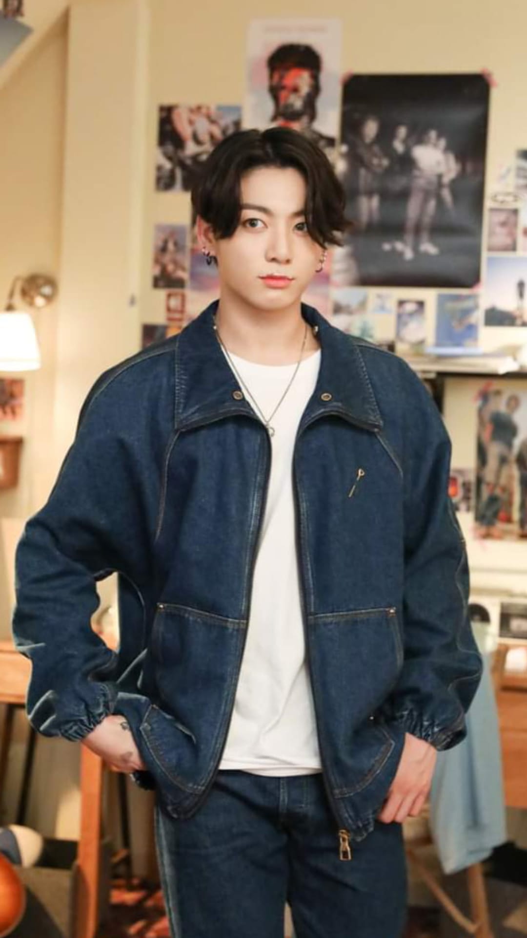 Jungkook wearing a denim jacket and jeans stands before a wall of posters in a warm, retro-style room.