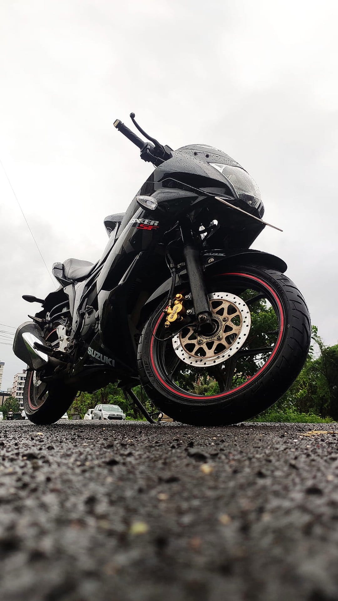 Suzuki Gixxer sportbike on wet asphalt with red rim accents, urban trees, and overcast sky in a low-angle view.