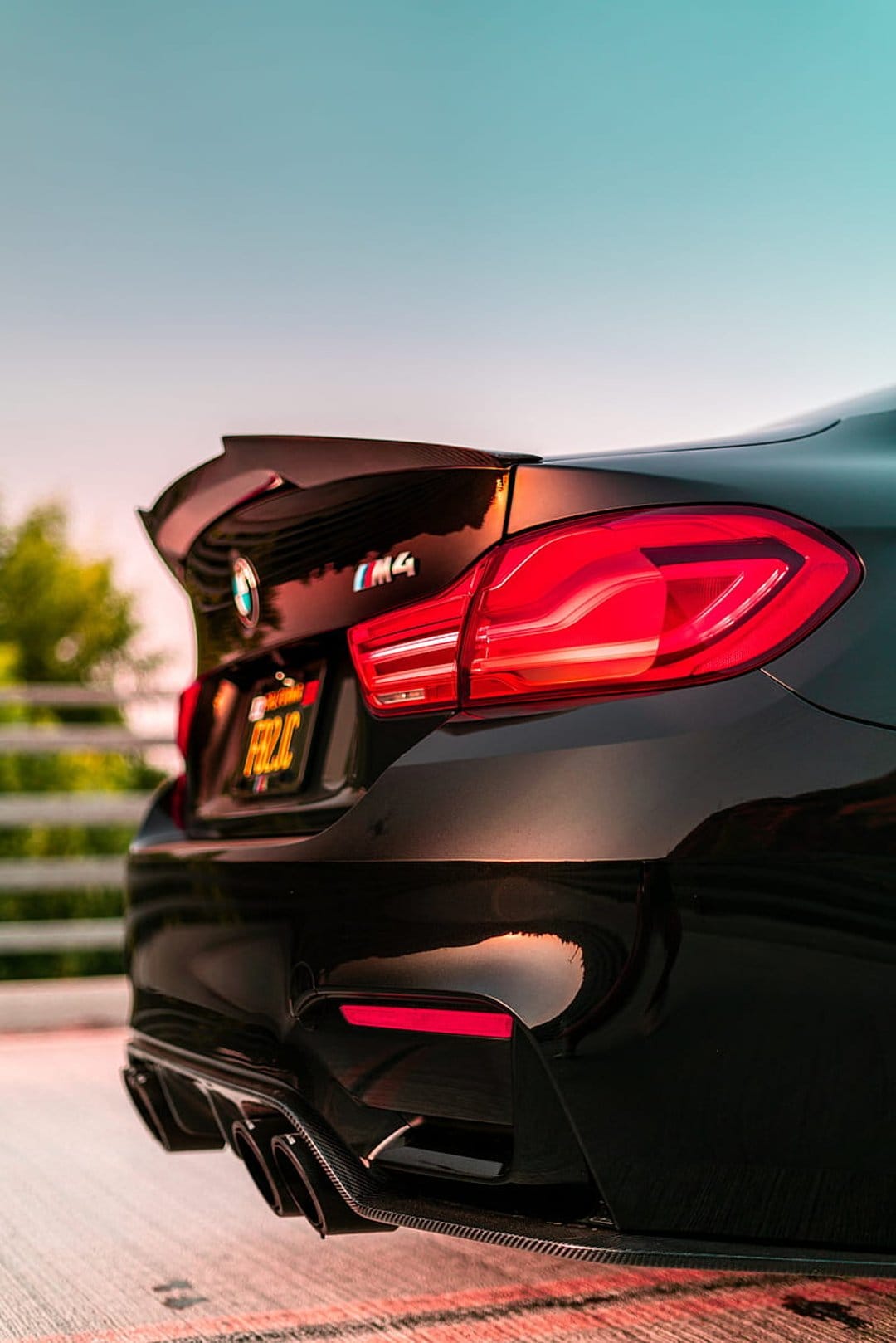 BMW M4 rear profile highlighting red LED tail lights, carbon fiber spoiler, and exhausts under sunset light.