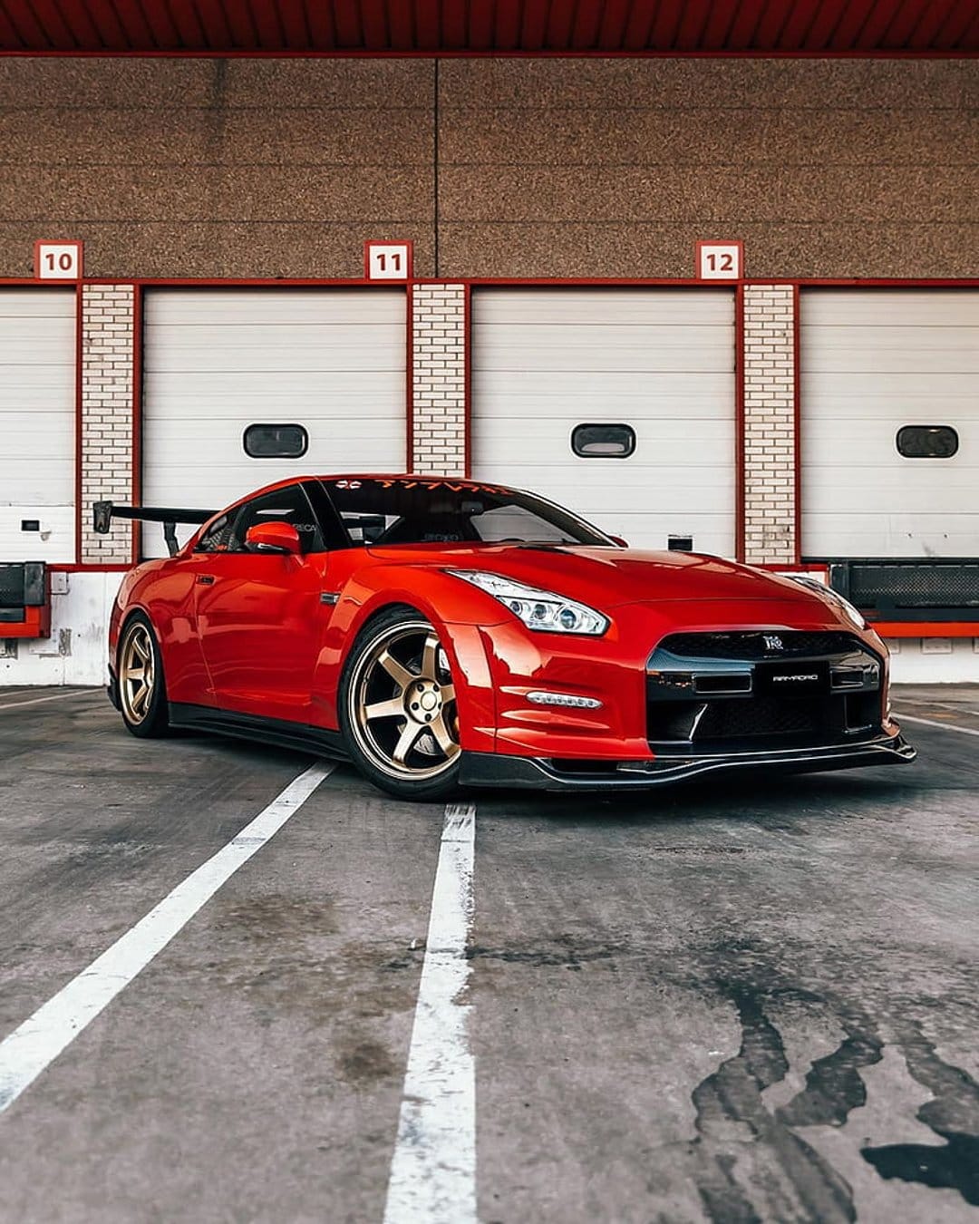 Red Nissan GT-R with gold wheels parked in front of industrial garage doors on a concrete lot with white lines.