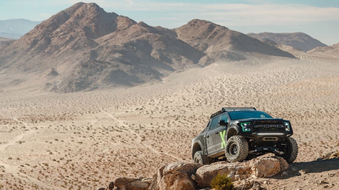 Black modified Ford Raptor truck perched on a rocky ledge overlooking a vast desert valley and mountains.