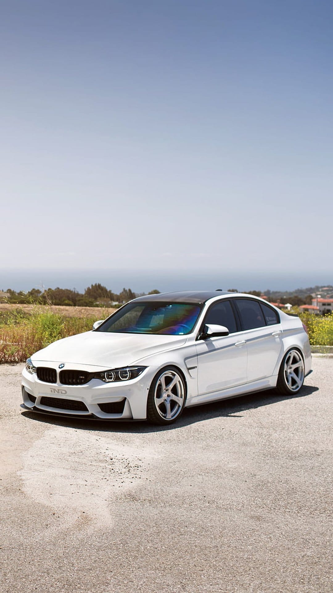 White BMW M4 Coupe with black wheels parked on a sunny coastal road overlooking the ocean and green hills.