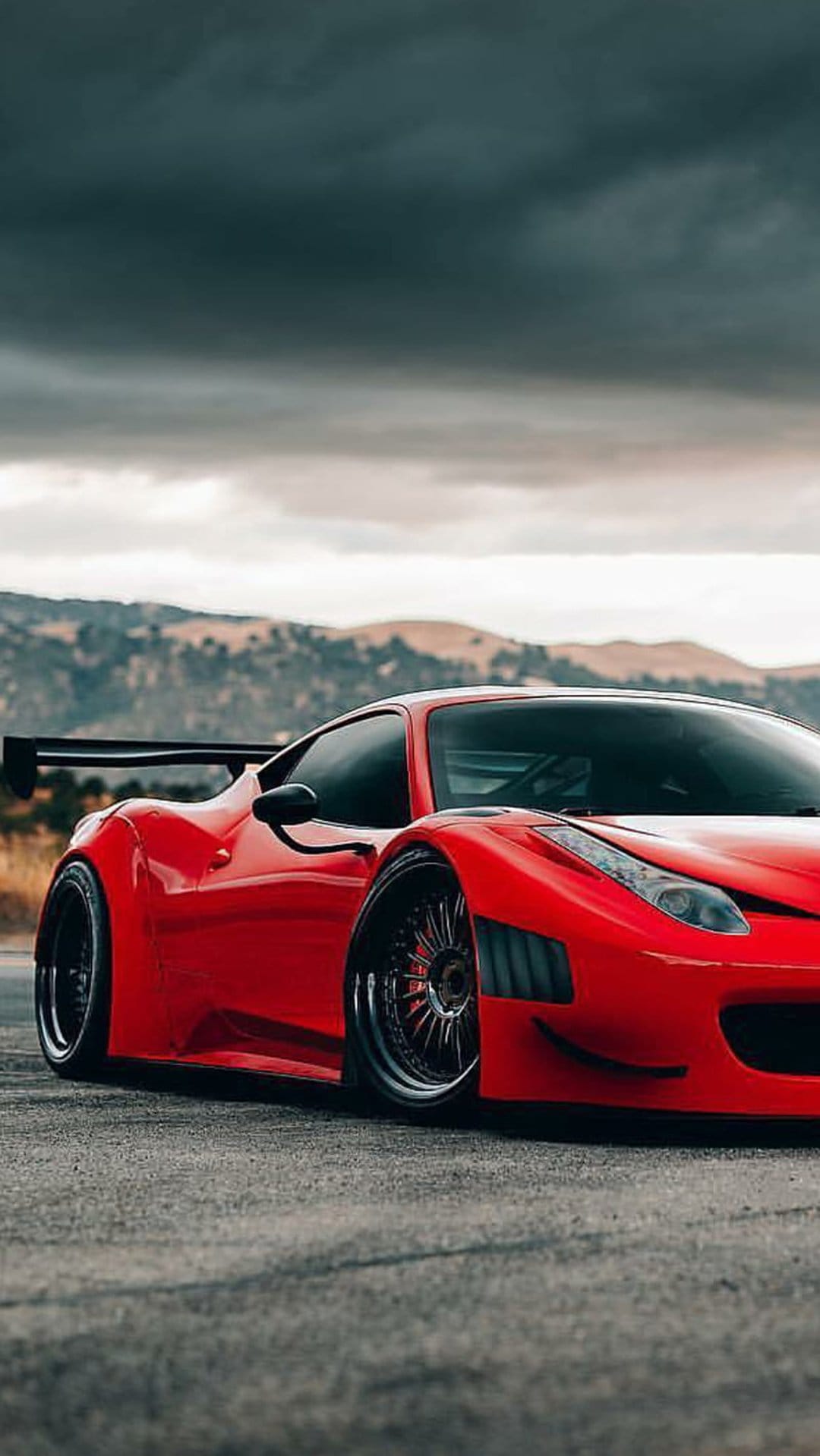 Red Ferrari 458 Italia track car with a large rear wing parked on a road against a dark, cloudy hill landscape.