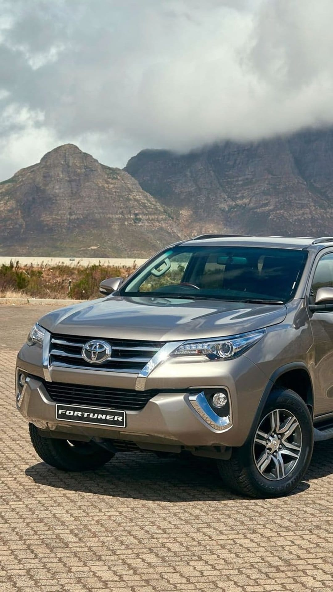 Tan Toyota Fortuner SUV parked on a paved road with sharp mountain peaks and a cloudy sky in the background.