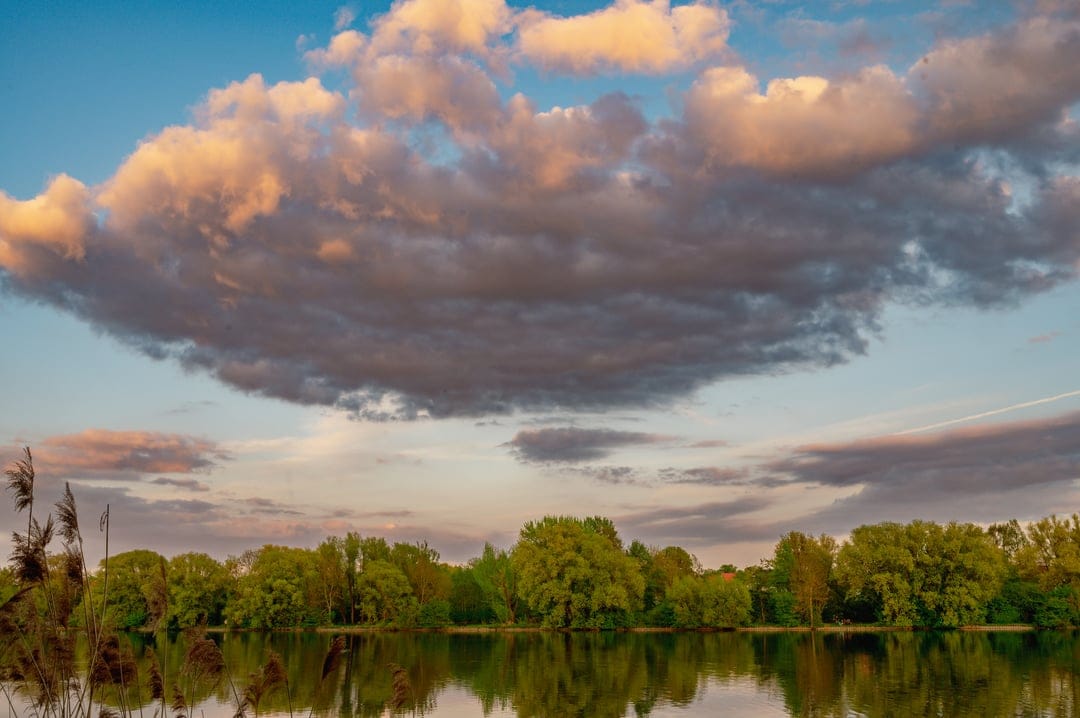 Sunset clouds in shades of orange and pink reflect on a mirror-like lake bordered by a dark silhouette of trees.