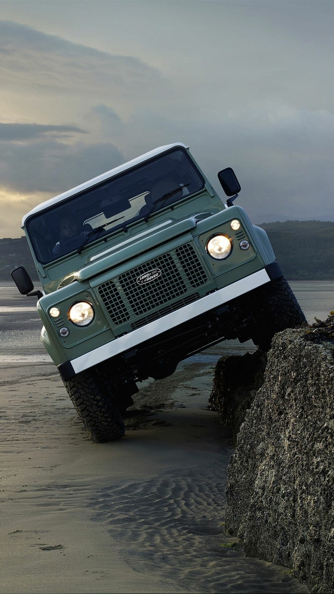 A sage green Land Rover Defender off-roading on a sandy beach, climbing a dark rock under an overcast sky.
