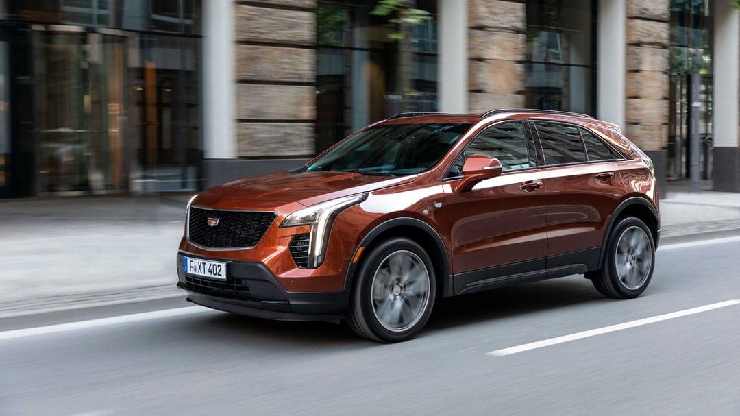 Bronze Cadillac SUV driving on a city street with motion blurred wheels and modern glass buildings behind.