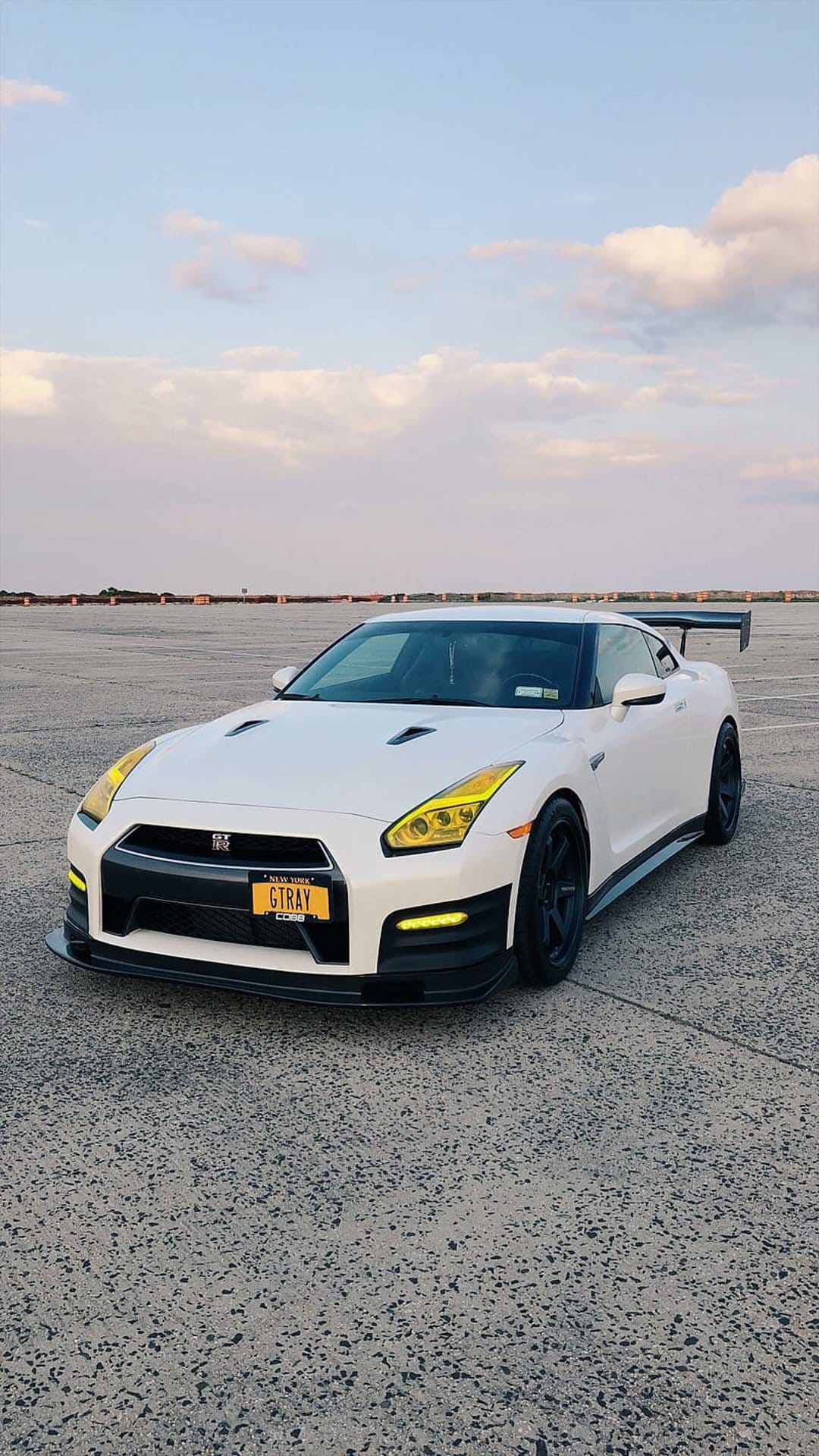 Nissan GT-R in white with yellow headlights and GTRAY license plate on asphalt under a cloudy blue sky.