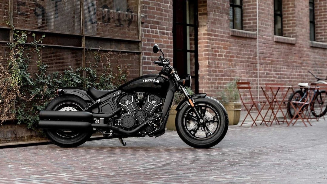 Black Indian Scout Bobber motorcycle parked on a brick courtyard beside an industrial red brick building.