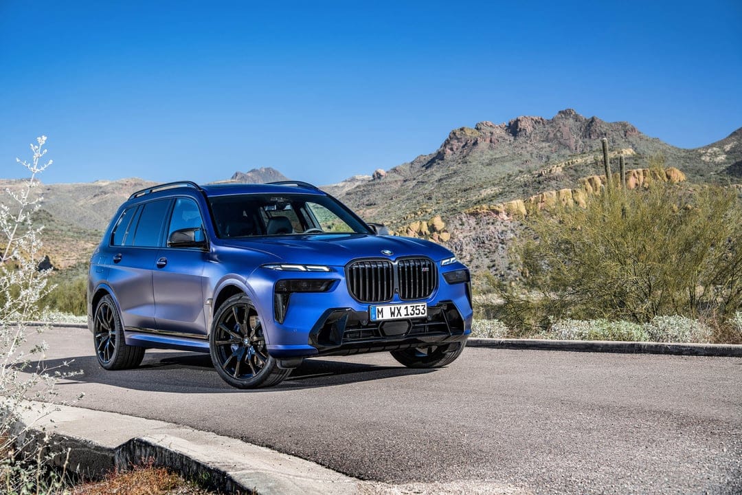 Matte blue BMW X7 M60i on a desert road with rugged mountains and a clear blue sky in the background.