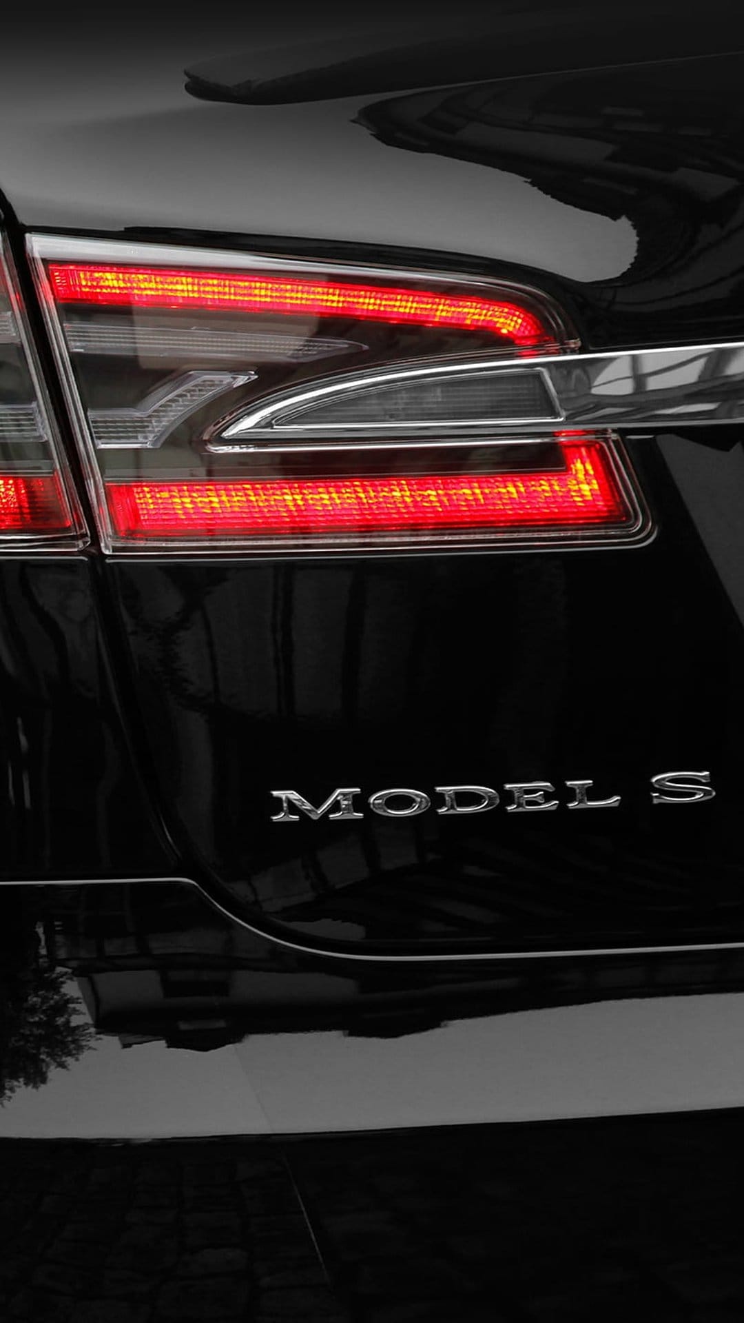 Tesla Model S rear red LED taillight and chrome badge on a polished black car body with ambient reflections.