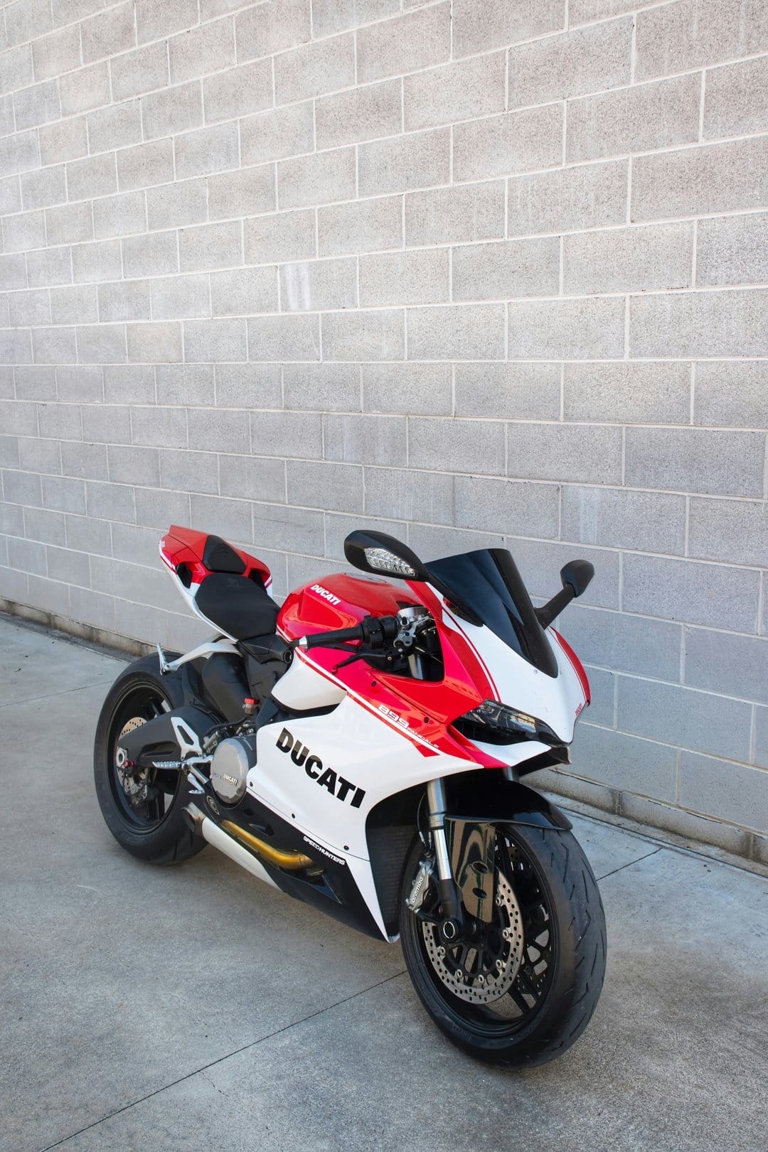 Red and white Ducati motorcycle parked beside a grey brick wall with visible disc brakes and tinted windscreen.