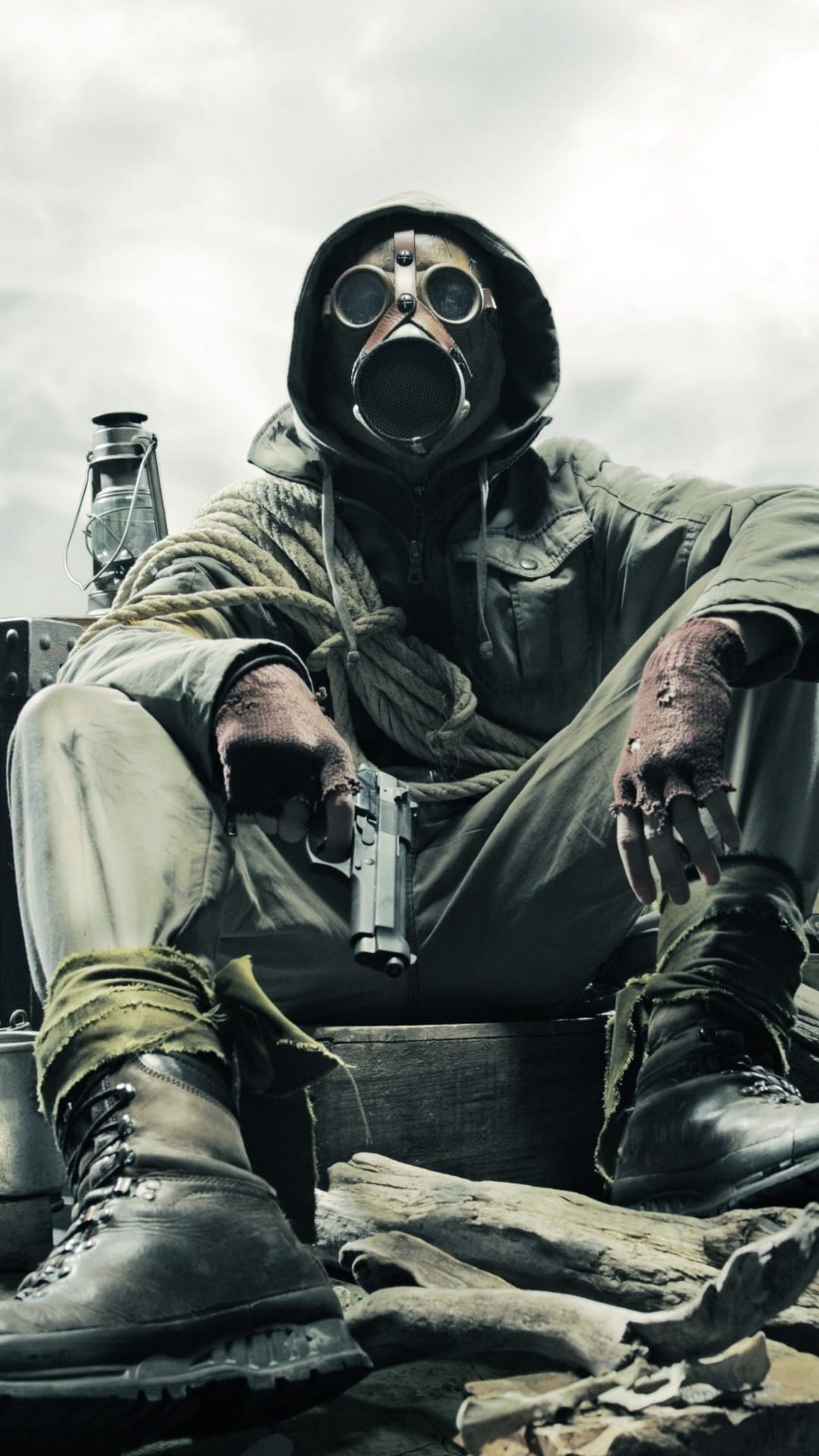 Survivalist wearing a gas mask and hood sits in a rocky wasteland next to an old lantern and coiled rope.