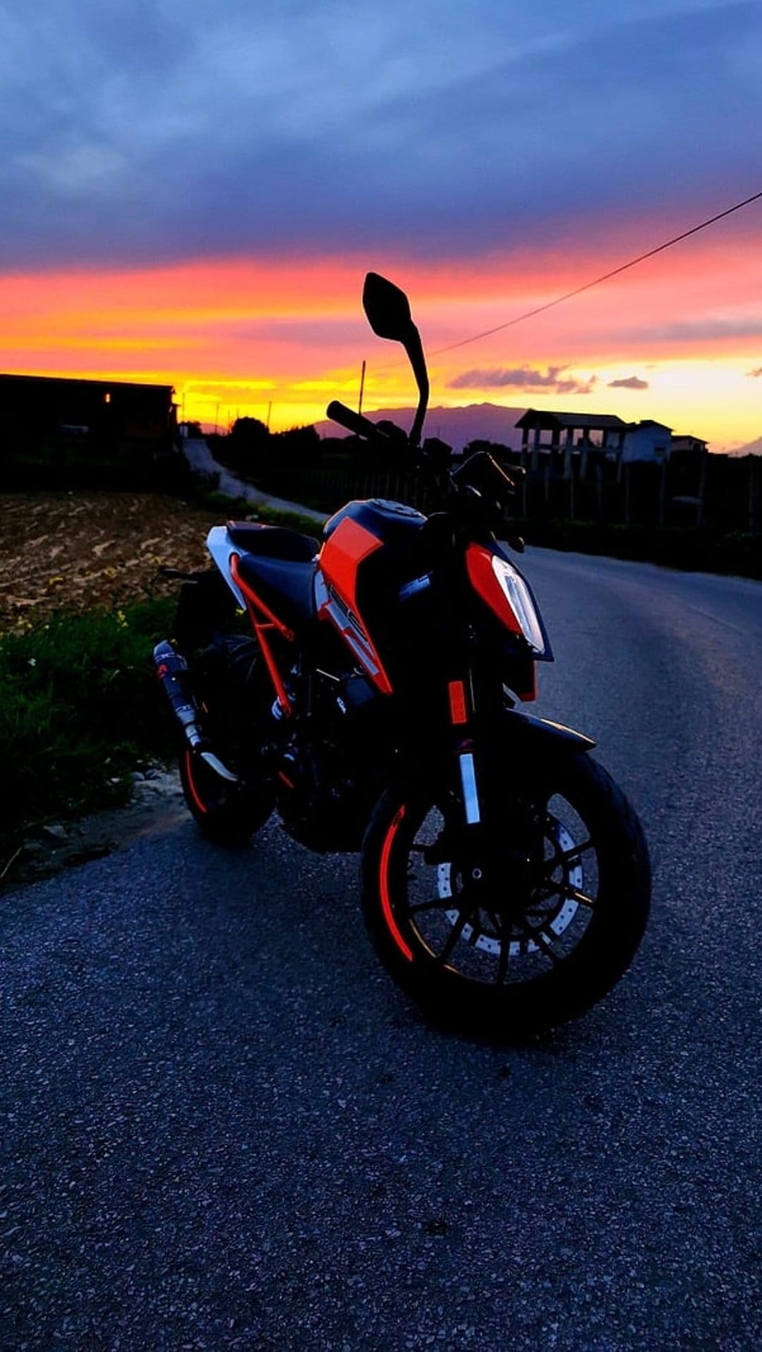 KTM motorcycle parked on a rural road under a glowing orange and purple sunset sky with distant farm fields.